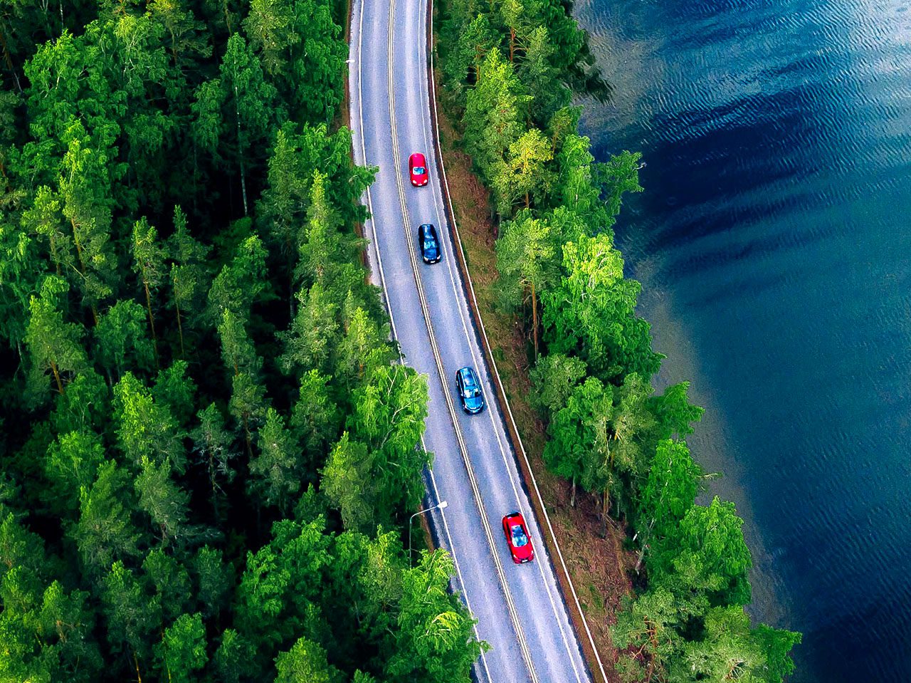Birds-eye aerial image of four cars, two blue and two red, driving along road beside lake. Trees on both sides of the road